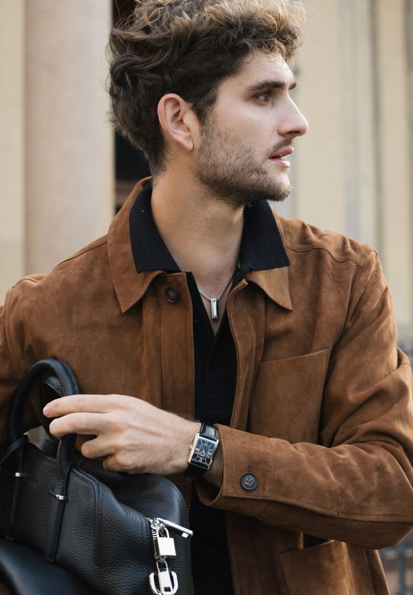 Brown suede jacket with contrasting black collar, holding a black leather bag. Features a silver watch and a plain black shirt.