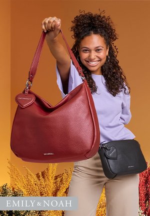 Sac de hobo en cuir rouge avec un accent en forme de cœur, associé à un petit sac bandoulière noir. L'arrière-plan présente un feuillage doré.