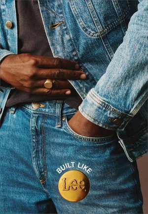 Jeansjacka i ljusblå nyans med kontrasterande gyllene detaljer, matchad med blå jeans som har en cirkulär gyllene patch med texten "Lee."