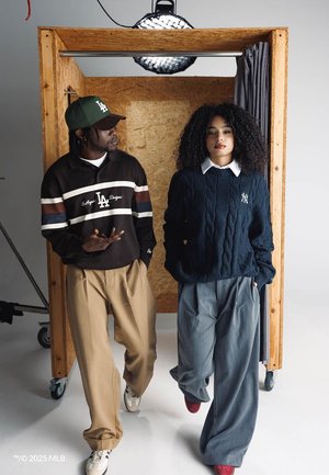 To personer iført løstsiddende bukser og MLB-holdtrøjer, stående inden for en træramme med en hængende studiolampe over.