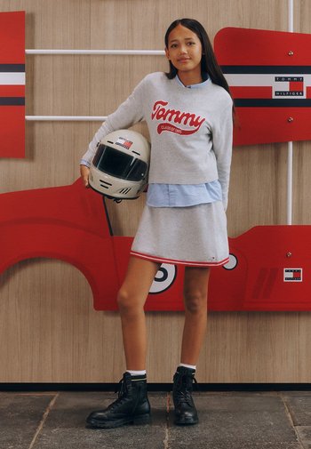 Grauer Pullover mit "Tommy"-Logo, über einem blauen Hemd getragen, kombiniert mit einem grauen Rock. Schwarze Stiefel und ein beiger Helm vervollständigen das Outfit.