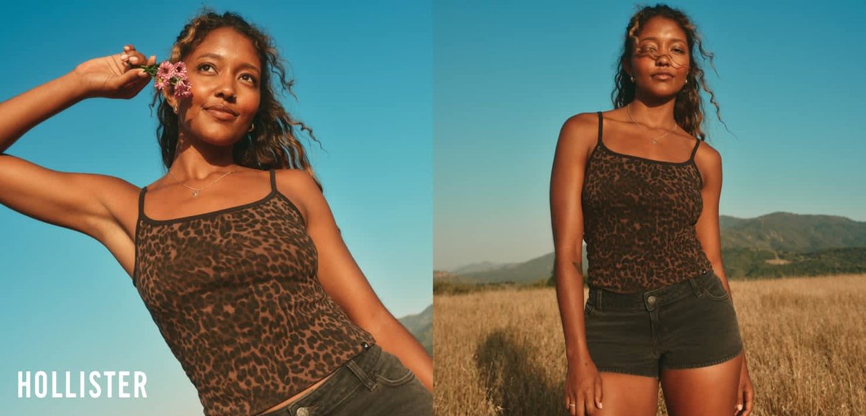 Frau in einem Tanktop mit Leopardenmuster und schwarzen Shorts posiert auf einem sonnigen Feld mit Bergen und klarem blauem Himmel im Hintergrund.