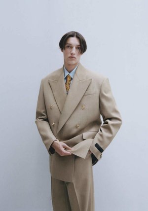 Beige double-breasted suit met gouden knopen en zwarte accenten. Getailleerde pasvorm, met notch-revers, gecombineerd met een gestreepte stropdas en een lichtblauw overhemd.