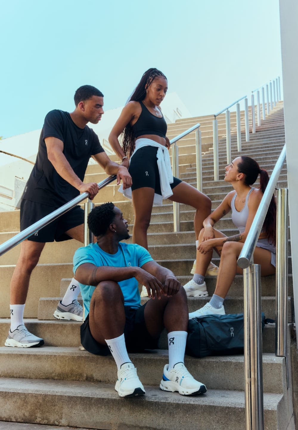 Vier Personen in Sportbekleidung sitzen und stehen auf Betontreppen. Schwarze Tanktops, bunte Shorts und Sportschuhe, mit einem Rucksack in der Nähe.