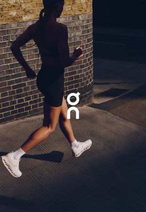 Runner in a black sports bra and shorts, wearing white athletic shoes with a distinctive design. Background features a brick wall.