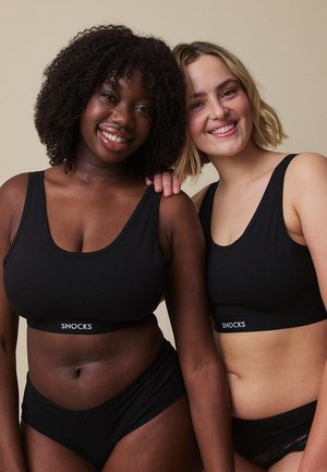To glade kvinder iført matchende sorte Snocks-braletter og undertøj, stående tæt sammen med den ene kvindes hånd på den andens skulder.