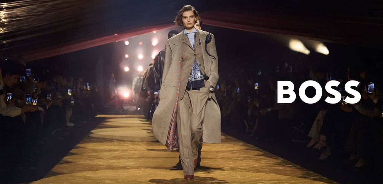 Modelka maszeruje po wybiegu w oversize'owym beżowym płaszczu i garniturze podczas pokazu mody, publiczność robi zdjęcia, po prawej stronie widoczne logo BOSS.