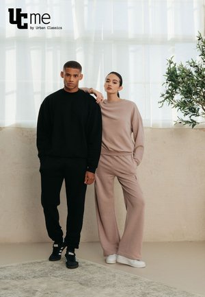Čierna voľná mikina a zodpovedajúce joggers na mužskom modeli; ružová voľná mikina a široké nohavice na ženskom modeli.