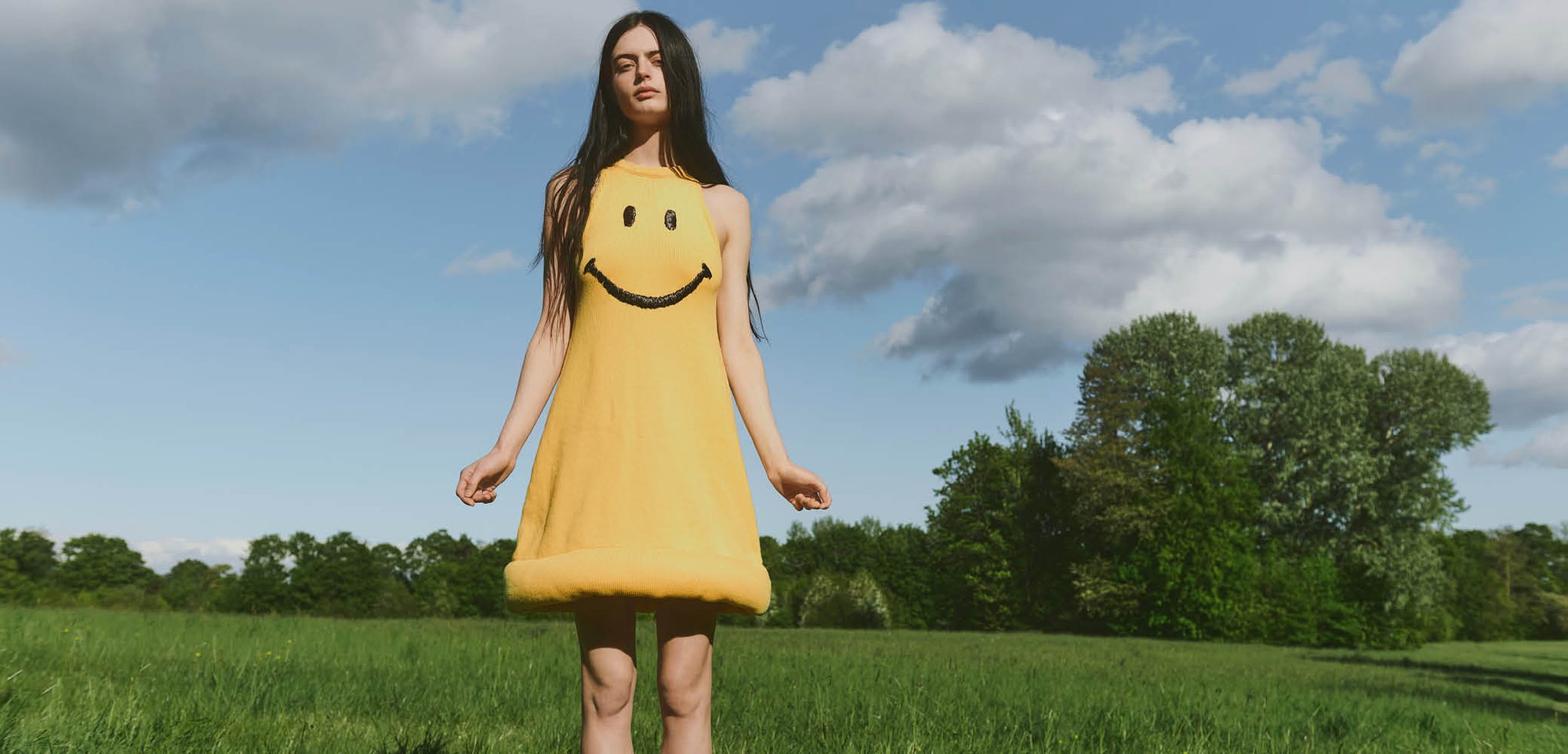 Ung kvinde står på en grøn mark iført en gul kjole med et sort smiley ansigt under en delvist overskyet blå himmel.