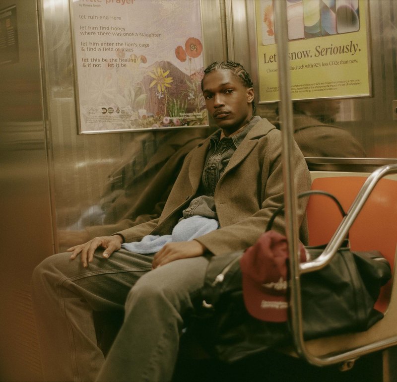 En person i en brun frakke sidder på et orange metro sæde iført en denimskjorte, grå bukser og en sort taske. Et lyseblåt håndklæde hviler på deres skød.