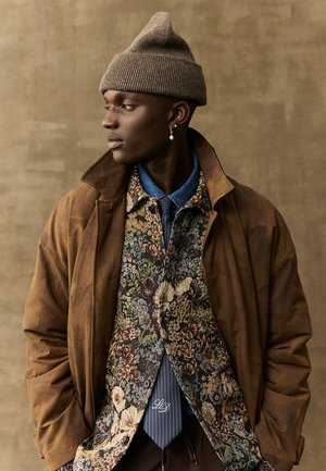 Bruine suède jack over een bloemenpatroon shirt met een blauwe denim kraag, een gestreepte stropdas, en een geribde bruine beanie. Perenvormige oorbel zichtbaar.