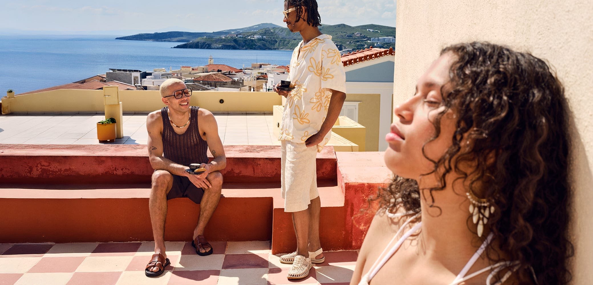 Troje ljudi na terasi s pogledom na more. Jedan nosi smeđi mrežasti top i šortseve, drugi ima cvjetnu košulju, a treći nosi kupaći kostim.