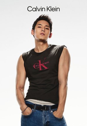 Black sleeveless t-shirt with red "CK" logo, paired with blue denim jeans. The model has short dark hair and is posing against a white background.