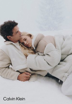 Dwóch osób siedzi na śniegu, ubranych w puszyste, kremowe i beżowe okrycia wierzchnie. Teksturowane materiały kontrastują z zaśnieżonym tłem.