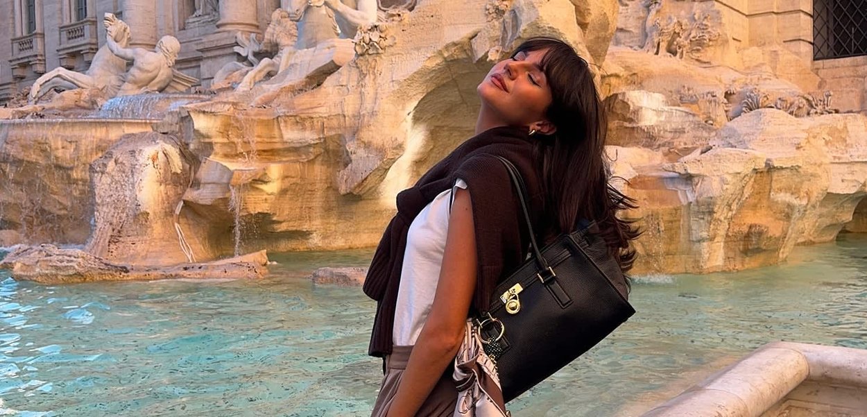 Donna con gli occhi chiusi inclinata all'indietro vicino a una grande fontana di pietra ornata con acqua che scorre e sculture, illuminata da una luce calda.