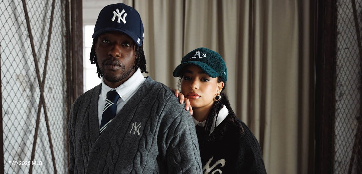 To personer iført MLB-holdkasketter og sweatere: mand med New York Yankees og kvinde med Oakland Athletics, poserer indendørs nær et vindue.