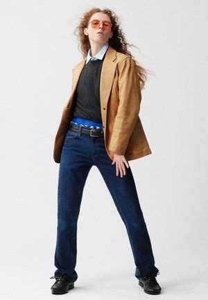 Beiger Wildlederblazer über einem grauen Pullover und blauem Hemd, kombiniert mit blauen Jeans und schwarzen Schuhen. Auffällige Details sind orangefarbene Sonnenbrillen und ein farbenfroher Gürtel.