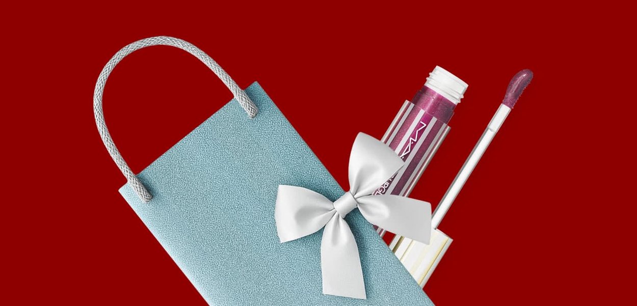 Una borsa regalo azzurro chiaro con una texture e un fiocco bianco contiene un gloss labbra lucido color bordeaux con un applicatore trasparente, su uno sfondo rosso.