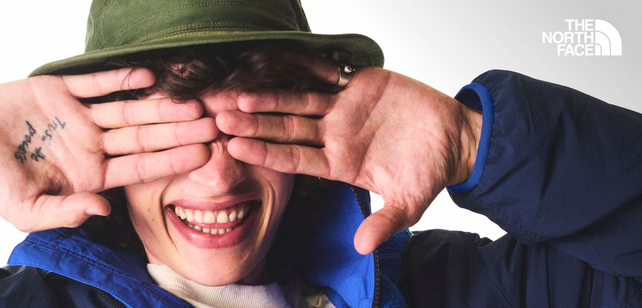 Person lächelt und bedeckt die Augen mit den Händen, trägt einen grünen Hut, eine blaue Jacke und ein weißes Hemd mit sichtbarem The North Face-Logo.