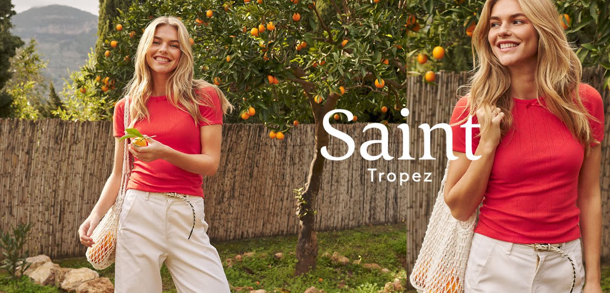 Lächelnde Frau in rotem Hemd und weißer Hose hält eine Netztasche mit Orangen, steht nahe Orangenbäumen und einem Holzzaun in einem sonnigen Garten.