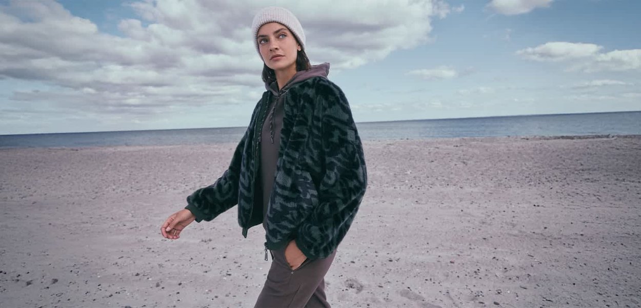 Sort og grøn mønstret faux fur jakke med hætte, parret med en grå sweatshirt og beige bukser, båret med en cremefarvet strikket beanie.