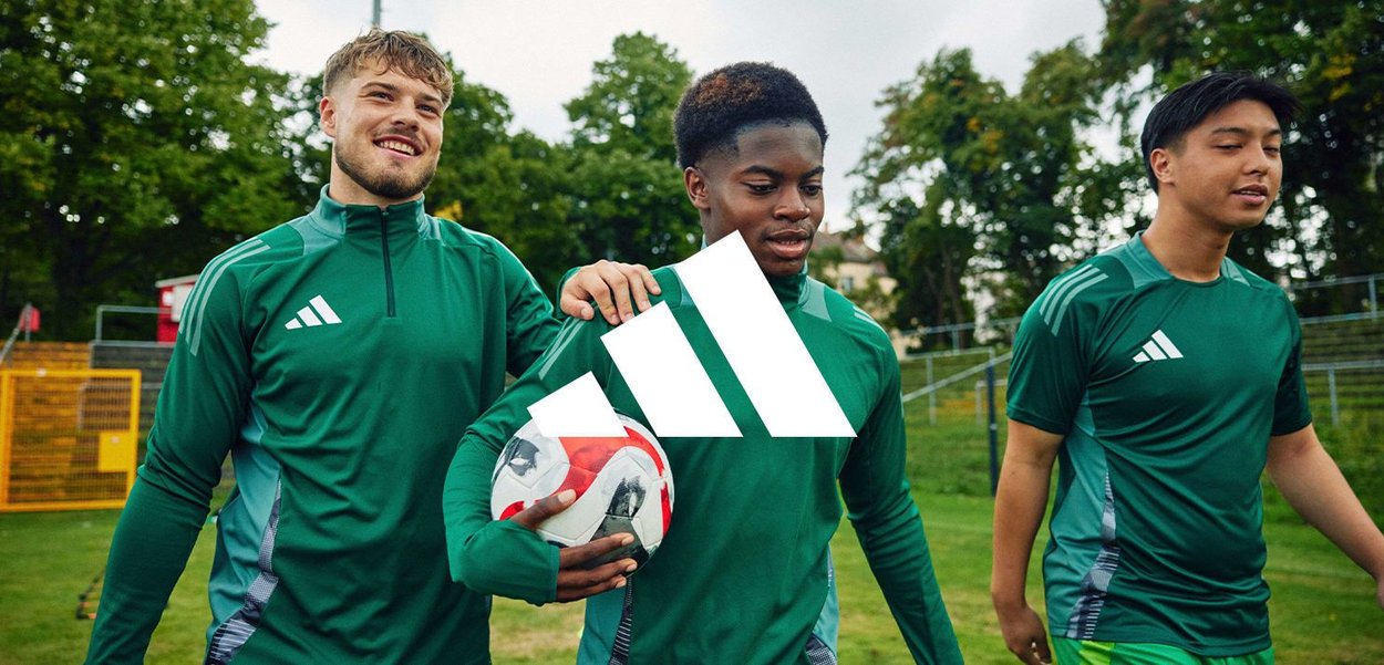 Trois jeunes hommes en tenue de sport verte marchent sur un champ herbeux, l'un d'eux tenant un ballon de football, entourés d'arbres et d'équipements de sport en plein air.
