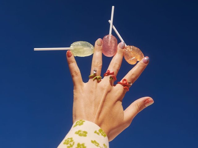 Πέντε πολύχρωμα λογαριάζι κρατιούνται από ένα χέρι στολισμένο με διάφορα δαχτυλίδια. Οι λογοριάζι είναι πράσινοι, ροζ και πορτοκαλί, ενώ τα δαχτυλίδια έχουν παιχνιδιάρικες σχεδιάσεις.