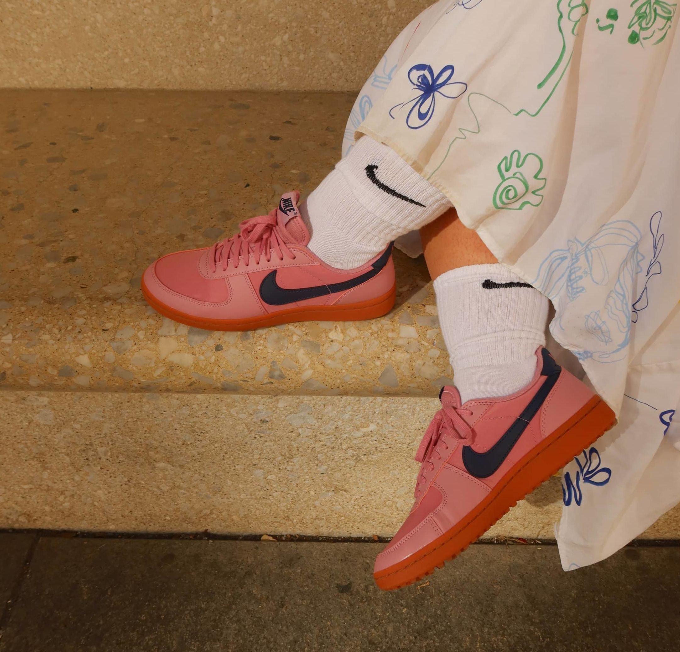 Pink Nike sneakers with darker purple accents, rubber sole, and matching laces; worn with white socks featuring a black logo.