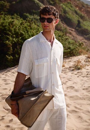 Junger Mann mit weißem kurzärmeligem Hemd und Sonnenbrille, der eine große beige Tasche trägt, steht am Sandstrand mit grünen Hügeln im Hintergrund.