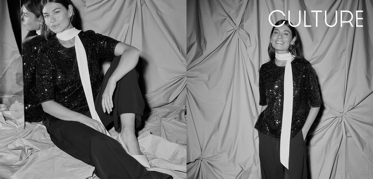 Haut noir à sequins avec manches courtes et une écharpe blanche, associé à un pantalon foncé. En arrière-plan, un tissu drapé et texturé.