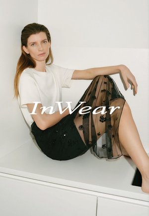 Schwarzer transparenter Rock mit floraler Stickerei, kombiniert mit einem weißen kurzärmeligen Oberteil. Das Model posiert sitzend vor einem neutralen Hintergrund.