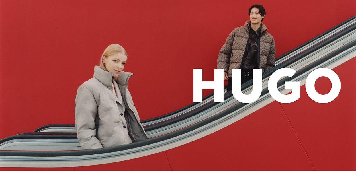 Chaqueta acolchada gris oversize en una mujer y chaqueta marrón acolchada en un hombre, ambos en una escalera mecánica frente a un fondo rojo brillante.