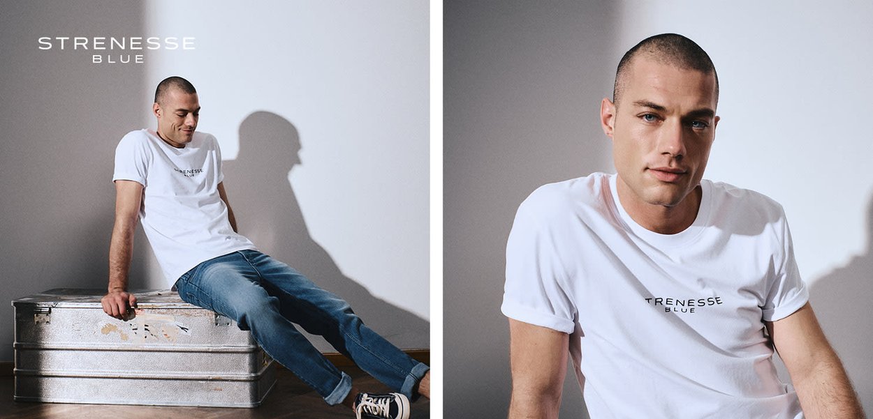Junger Mann trägt ein weißes Strenesse Blue T-Shirt und Jeans, sitzt auf einem metallischen Koffer vor einem schlichten hellen Hintergrund.