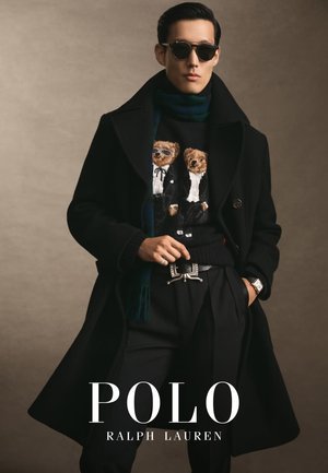 Zwarte overjas gedragen over een zwarte trui met twee beren, zwarte broek en een zilveren ceintuur. Accessoires zijn onder andere zonnebril en een horloge.