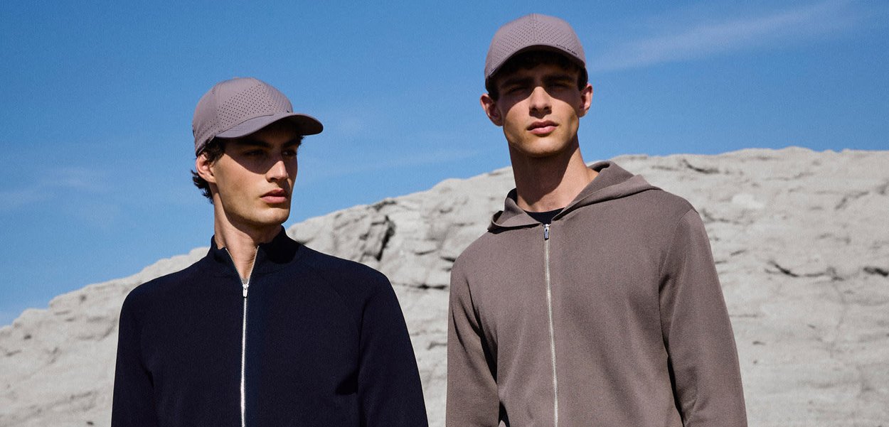 Zwei Männer, die neutrale Reißverschlussjacken und hellgraue Kappen tragen, stehen im Freien vor felsigem Gelände und klarem Himmel im Hintergrund.