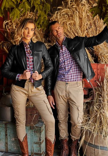 Chaquetas de cuero negras, camisas de cuadros, pantalones cargo beige y botas de vaquero marrones. Fondo texturizado con heno y elementos de madera.