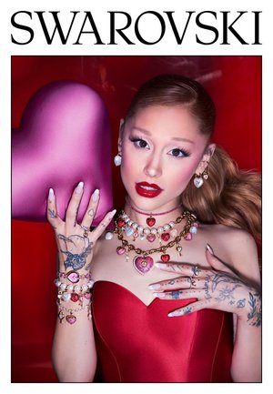 Donna con rossetto rosso e tatuaggi indossa gioielli a tema cuore e un vestito rosso, tenendo un grande cuore rosa su uno sfondo rosso.