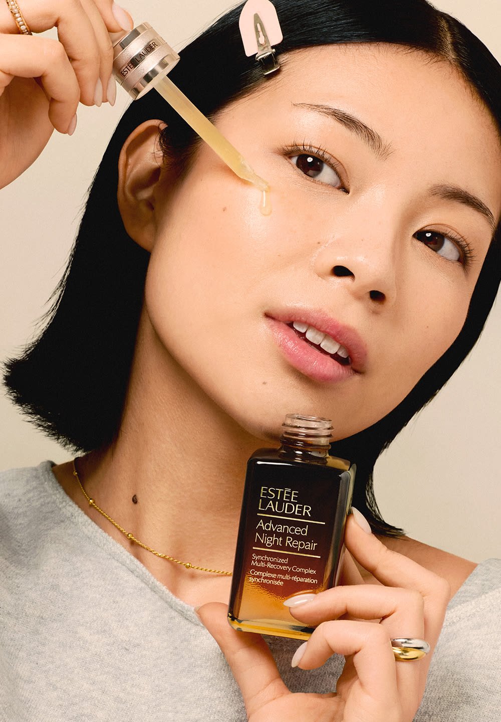 Vrouw die Estée Lauder Advanced Night Repair serum met een druppelaar op haar wang aanbrengt, die haar huidverzorgingsroutine en productdetails toont.