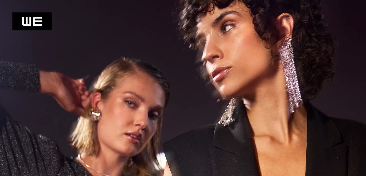 Twee modebewuste vrouwen die sprankelende oorbellen en zwarte outfits dragen, poseren tegen een donkere achtergrond met het "WE"-logo in de hoek.