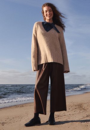 Lächelnde Frau steht an einem sandigen Strand, trägt einen beigen Pullover, dunkle, weit geschnittene Hosen, schwarze Schuhe und einen dunkelblauen Schal mit einer Schleife.