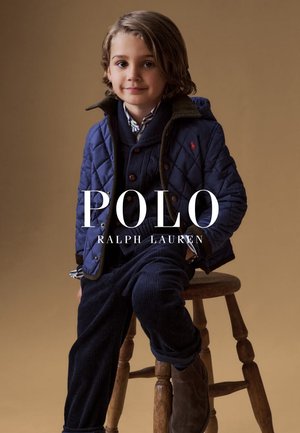 Ragazzo giovane seduto su uno sgabello di legno che indossa una giacca trapuntata blu navy, un maglione a maglia, una camicia a righe, pantaloni in velluto a coste e stivali marroni su sfondo beige.