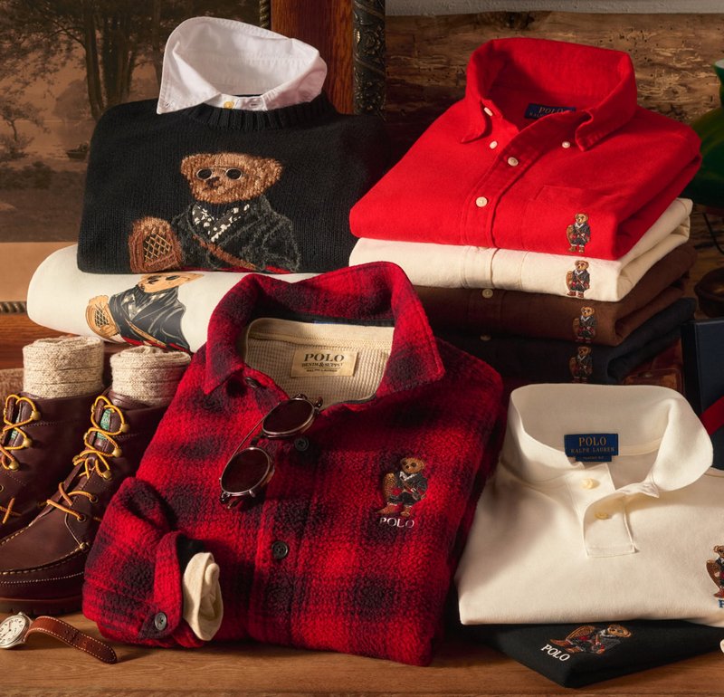 Exhibición de ropa Polo Ralph Lauren: suéter negro, camisa de franela roja, camisetas variadas, botas marrones y accesorios sobre fondo de madera.