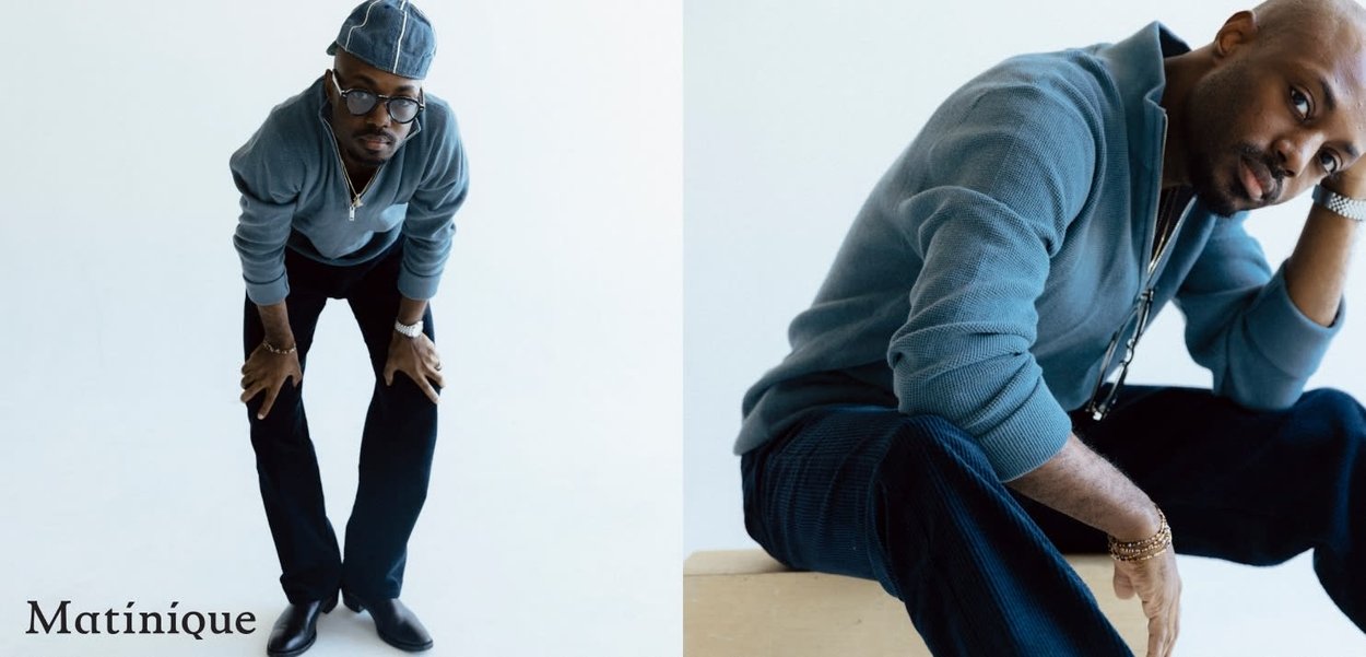 Lässiges Outfit mit einem blauen Strickpullover und dunklen weiten Hosen, runden Brillen und einer Mütze, in einem hellen Umfeld posierend.