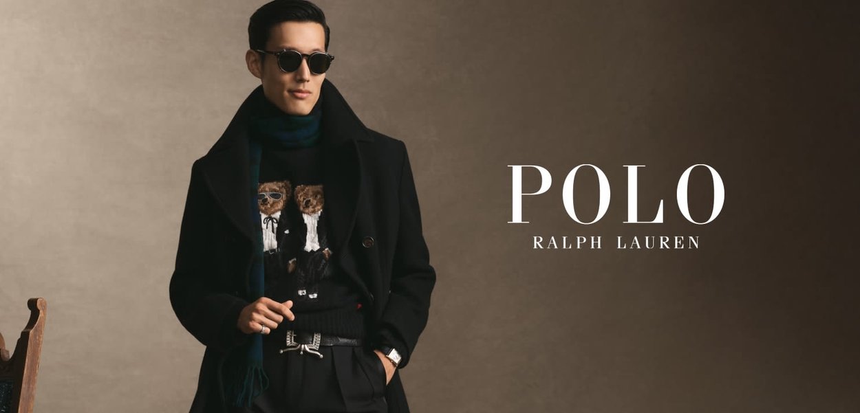 Schwarzer doppelt-reihiger Mantel über einem gemusterten Pullover mit Bärendesigns, kombiniert mit Sonnenbrille und einem grünen Schal. Dunkler Hintergrund.