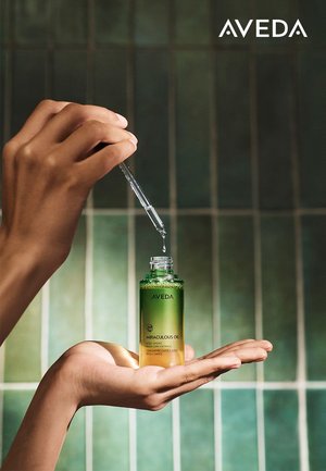 Aveda Miraculous Oil-flesje met een groene en gouden gradient, voorzien van een pipet. Een hand houdt het flesje vast tegen een groene tegels achtergrond.