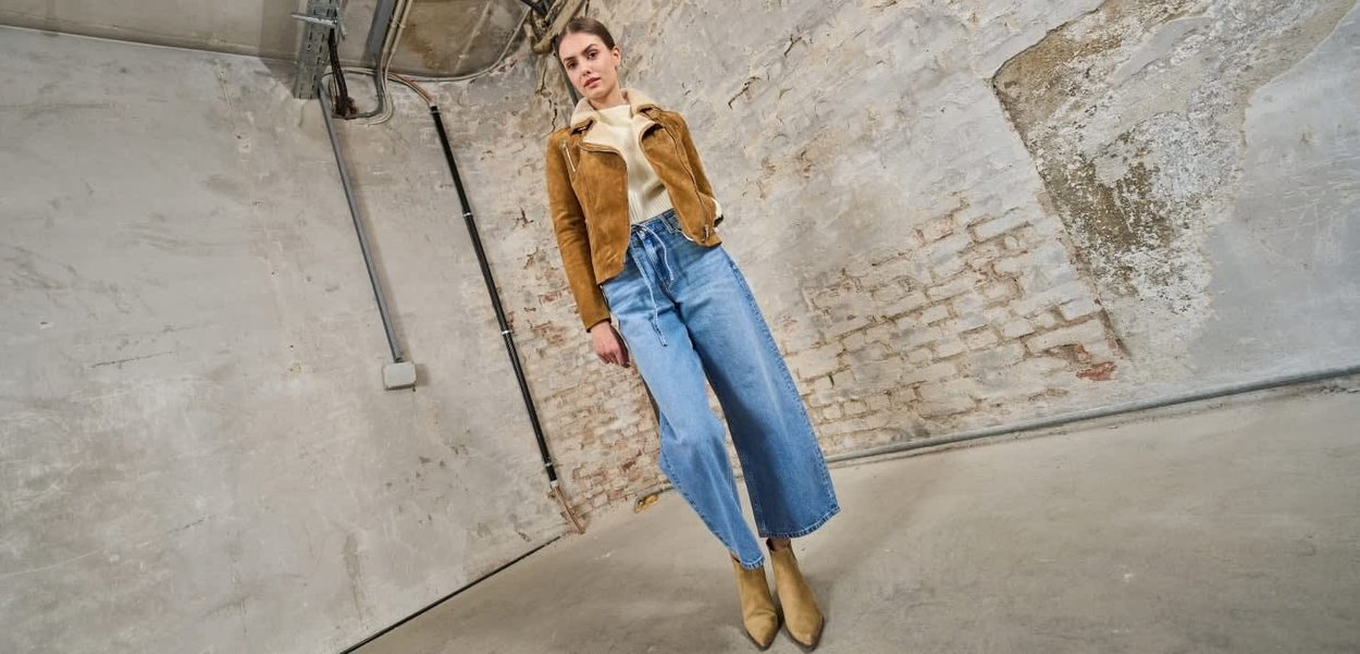 Brązowa zamszowa kurtka, kremowy sweter, dżinsy o wysokim stanie i szerokich nogawkach oraz beżowe botki na tle fakturowanej ściany.