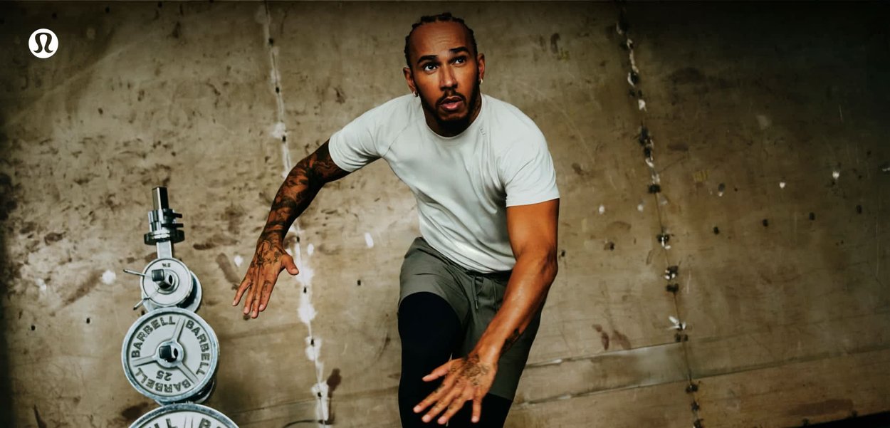 Athlet in weißem Hemd und grauer Shorts führt eine dynamische Übung im Fitnessstudio aus, neben gestapelten Langhantelgewichten vor einer abgenutzten Wand.