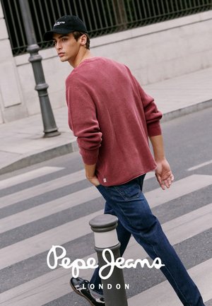 Joven con gorra negra, suéter rojo y vaqueros azules cruzando una calle de la ciudad cerca de un bolardo peatonal.