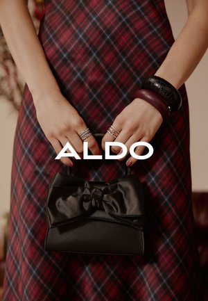 Zwarte handtas met een grote satijnen strik, vastgehouden in handen versierd met meerdere ringen, tegen een rood en blauw geruit patroon jurk.