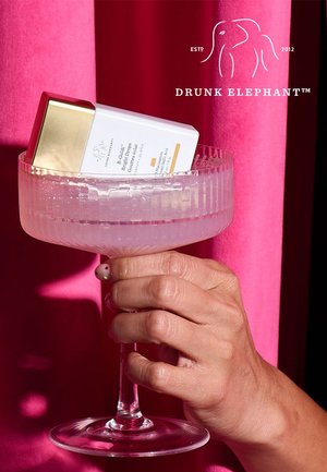 Drunk Elephant product in a clear, ribbed glass, held by a hand. The box is white with a gold cap, featuring orange text and details.